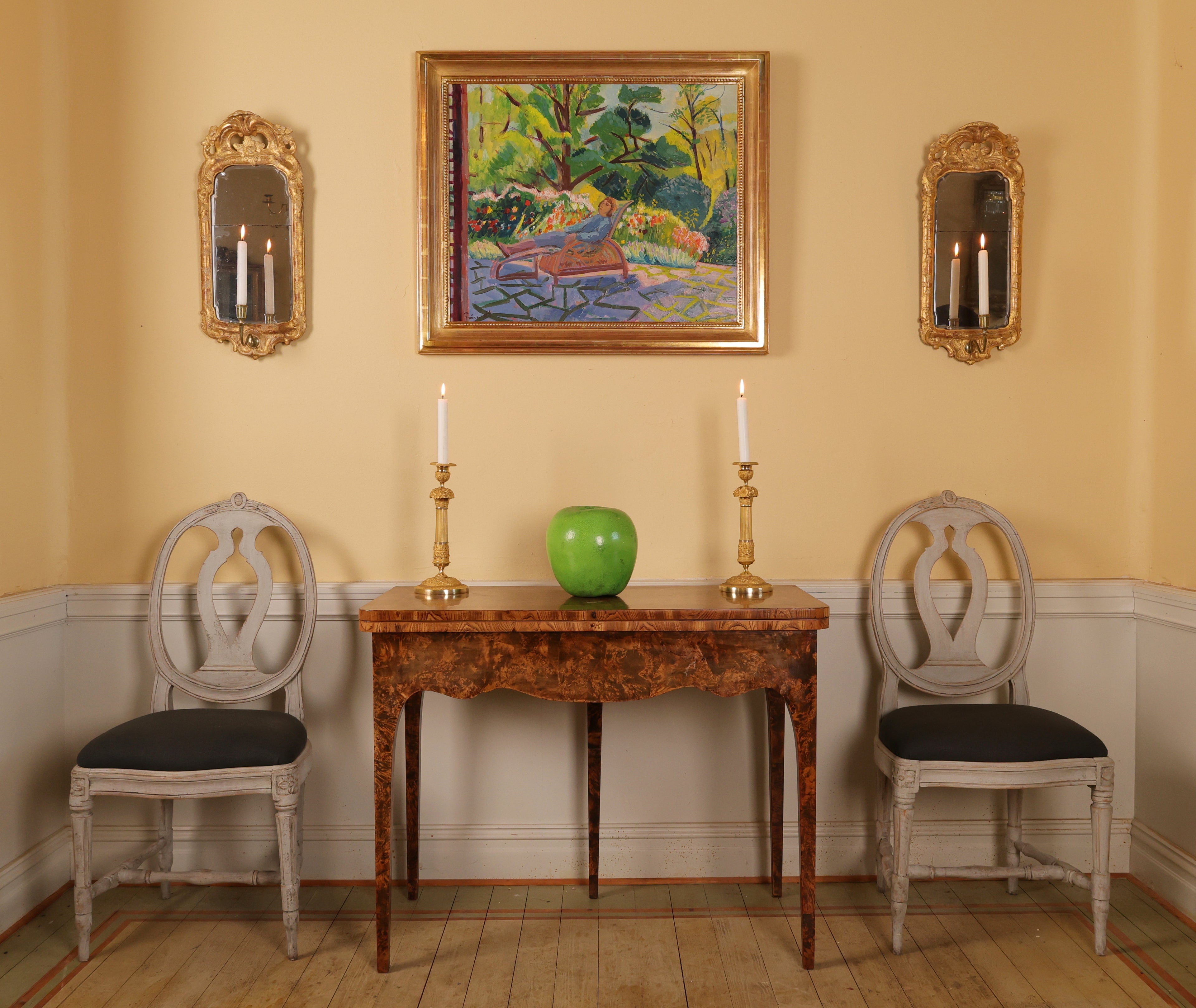 Banner Image of Napoleon Antique & Art, depicting a Green Apple with two Empire Candlesticks with Garlands on a brown antique table. On each side of the table are two grey antique chairs with black fabric. Above the table is a lush Isaac Grünewald Painting depicting a person laying on a sunbed in a garden with flowers and trees in the background. On the sides of the paining hang two gilded mirros with lit candles.