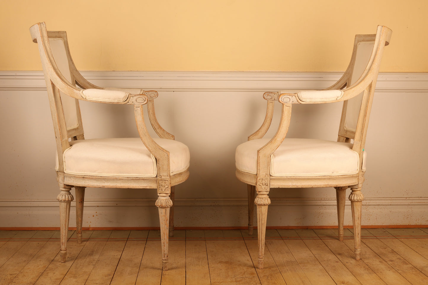 Side view of A pair of pearl-grey Gustavian antique armchairs from the late 18th century with white upholstery. The chairs feature carved leaf motifs and stand on collared, fluted legs.