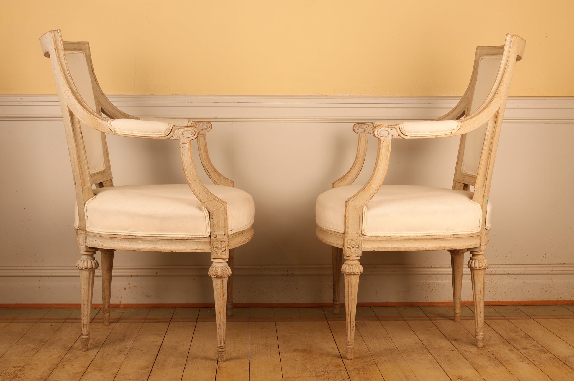 Side view of A pair of pearl-grey Gustavian antique armchairs from the late 18th century with white upholstery. The chairs feature carved leaf motifs and stand on collared, fluted legs.