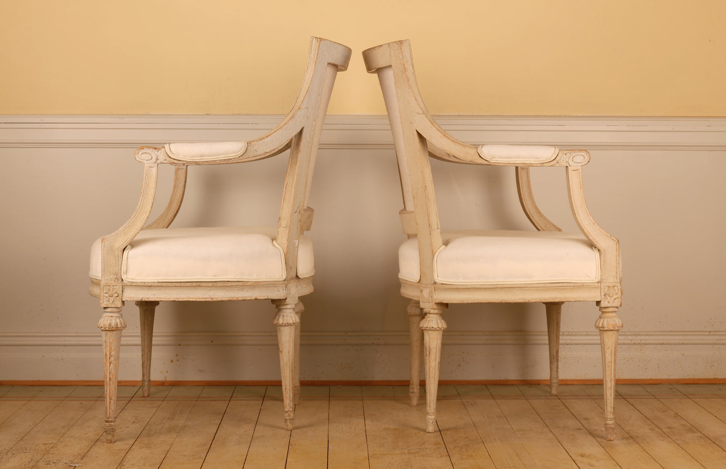 Side view of A pair of pearl-grey Gustavian armchairs from the late 18th century with white upholstery. The chairs feature carved leaf motifs and stand on collared, fluted legs.