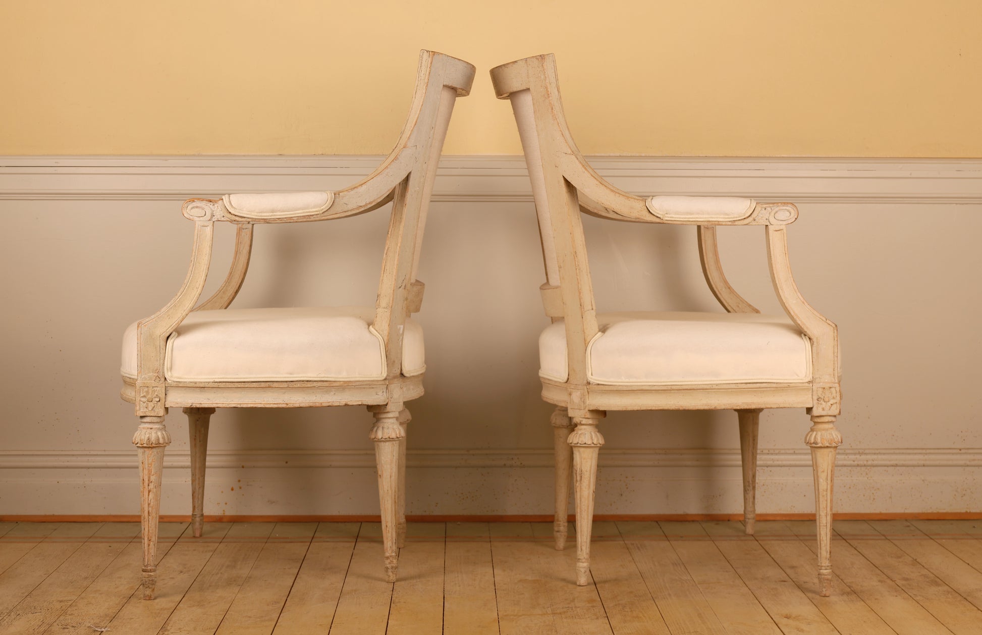 Side view of A pair of pearl-grey Gustavian armchairs from the late 18th century with white upholstery. The chairs feature carved leaf motifs and stand on collared, fluted legs.