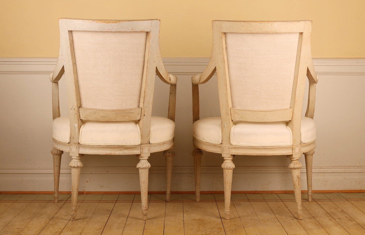 Backview of a pair of pearl-grey Gustavian armchairs from the late 18th century with white upholstery. The chairs feature carved leaf motifs and stand on collared, fluted legs.