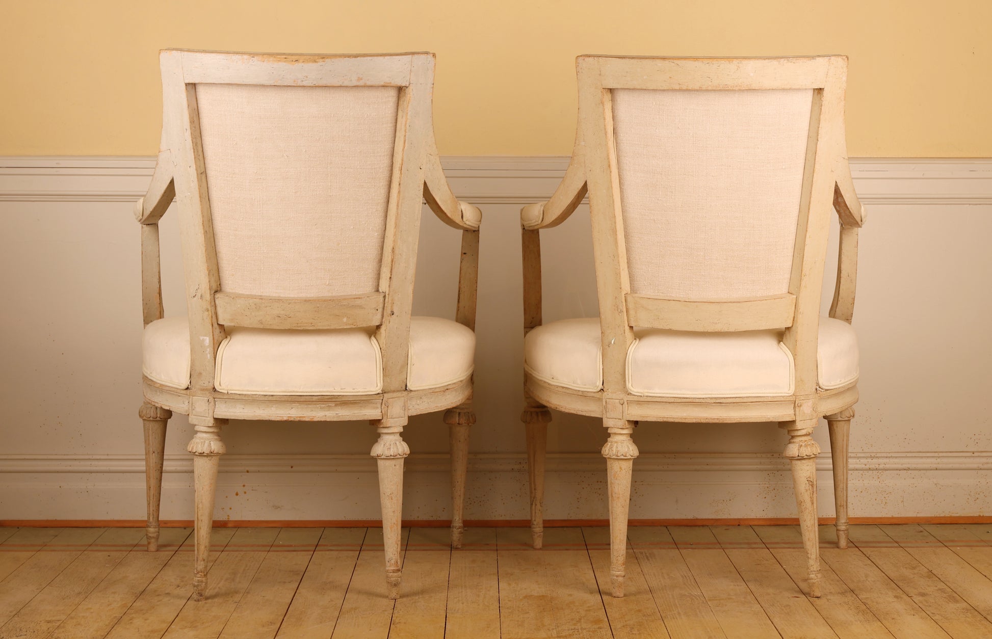 Backview of a pair of pearl-grey Gustavian armchairs from the late 18th century with white upholstery. The chairs feature carved leaf motifs and stand on collared, fluted legs.