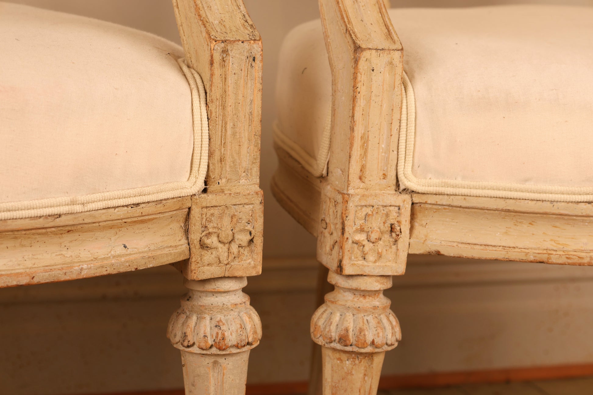 Close up of A pair of pearl-grey Gustavian armchairs from the late 18th century with white upholstery. The chairs feature carved leaf motifs and stand on collared, fluted legs.