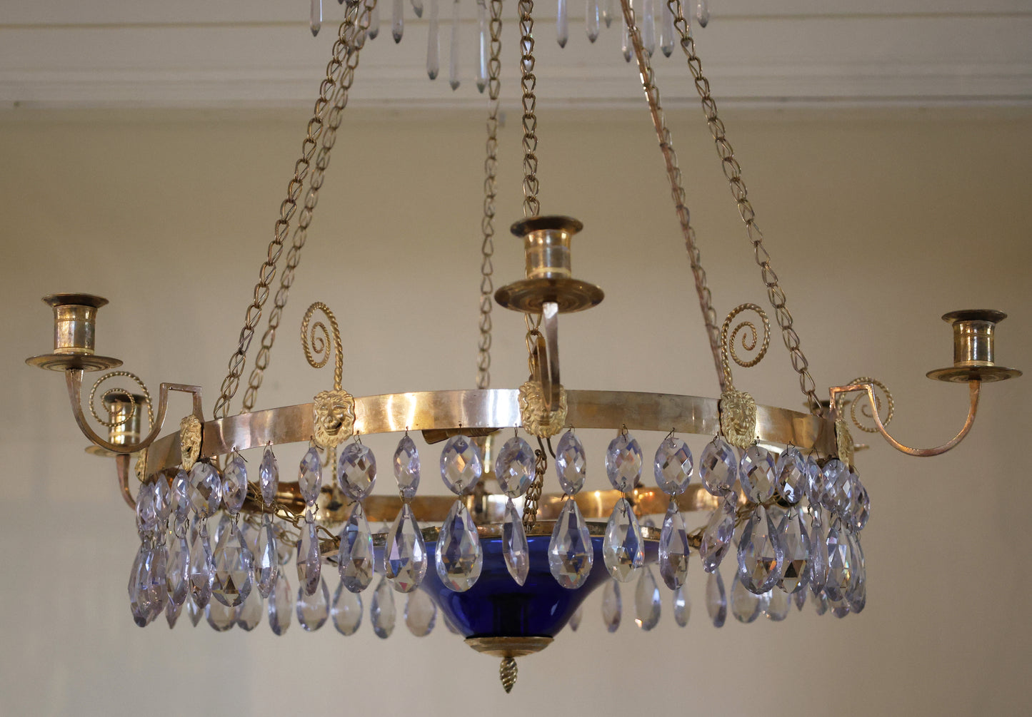 A late-Gustavian chandelier with a blue bowl, made in Stockholm in the 1790s. The burnished gilded central ring is decorated with lion mask carvings. The ring features six light arms, and six chains hold the ring together with the upper part, which consists of a "bishop's mitre" with a plume. The blue glass bowl contains a candle holder for a small candle.