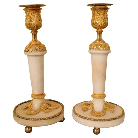 A pair of French Louis XVI candlesticks from the late 18th century. The stems and bases are made of white Carrara marble, while the fittings are burnished gilded bronze. The base is decorated with drapery and tassels, and the light sockets feature acanthus leaf decorations. The candlesticks rest on three ball feet. White background.