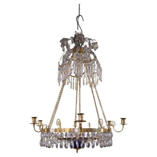 A late-Gustavian chandelier with a blue bowl, made in Stockholm in the 1790s. The burnished gilded central ring is decorated with lion mask carvings. The ring features six light arms, and six chains hold the ring together with the upper part, which consists of a "bishop's mitre" with a plume. The blue glass bowl contains a candle holder for a small candle. White background.