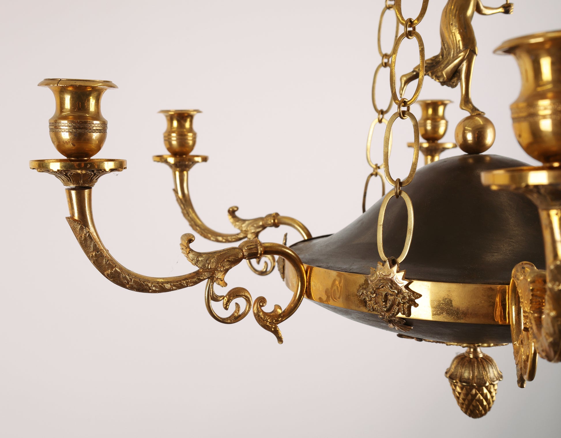 Details of High-class Empire pendant from Stockholm around 1800-1810. It features six beautifully decorated candle arms with acanthus leaves. The central decoration includes a female figure standing on a sphere. Three chains support the pendant, which culminates at the top with a plate before the ceiling hook. The ring is adorned with Medusa heads. The pendant is finished at the bottom with a decoration of a pine cone in fire-gilded bronze.